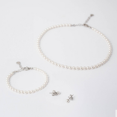 Esta é uma coleção de acessórios de prata banhada a ródio com pérolas Murano, exibida contra um fundo branco. O conjunto inclui um colar e uma pulseira, ambos apresentando uma única fileira de pérolas brancas lustrosas espaçadas uniformemente. Acompanhando estes estão um par de brincos, cada um consistindo de uma única pérola Murano em prata banhada a ródio. A simplicidade e elegância do design destacam a beleza das pérolas e o acabamento metálico brilhante.