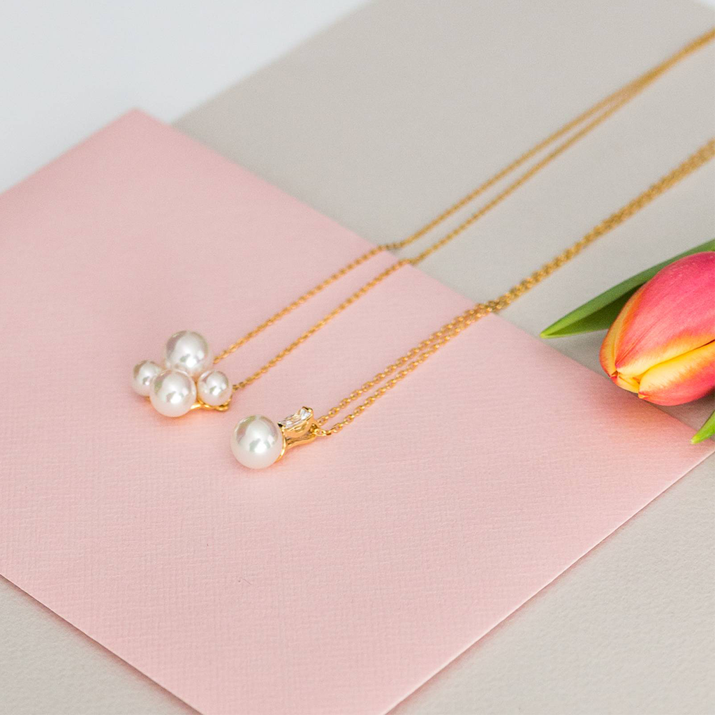 Este é um colar de prata banhado a ródio, exibido numa superfície rosa pastel ao lado de uma tulipa com a sua folha verde parcialmente visível. O colar apresenta uma corrente delicada e um pendente adornado com pedras de zircónia cintilantes, que refletem a luz e adicionam elegância à peça. A imagem destaca o design do colar e o contraste do brilho metálico contra o fundo suave, enfatizando a sua beleza e artesanato.