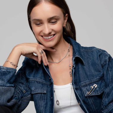 Mulher com casaco de ganga sobre uma blusa branca, adornada com várias peças de joalharia, incluindo colares, pulseiras e um anel.