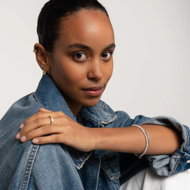 imagem de mulher de casaco jeans sobre uma blusa branca. A pessoa tem um brinco na orelha visível e está adornada com um anel no dedo anelar da mão esquerda e uma pulseira no pulso esquerdo. A mão está colocada suavemente no ombro, sugerindo uma pose relaxada ou casual.