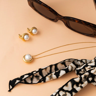 Colar de prata banhada a ródio com um pendente de pérola Murano. O colar é exibido sobre um fundo pastel, acompanhado por brincos combinando com molduras douradas e um par de óculos de sol com armação tartaruga. Um lenço preto e branco com padrão adiciona um toque elegante à composição, destacando a beleza refinada e o artesanato do colar.
