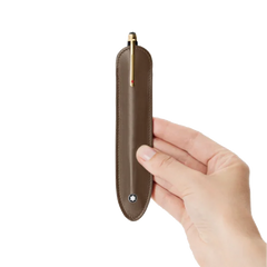 Uma mão segura uma elegante capa de caneta esferográfica castanha com detalhes dourados e um clipe, embainhada em um estojo de couro castanho com costura visível e fecho prateado