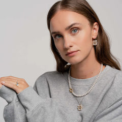 Pessoa usando um colar de prata banhada a ouro com pérolas de concha. O colar tem um design clássico com pérolas espaçadas uniformemente e um pendente central maior, possivelmente uma pérola ou motivo floral. A pessoa também usa brincos e anel combinando, sugerindo um conjunto de joias elegante e coordenado