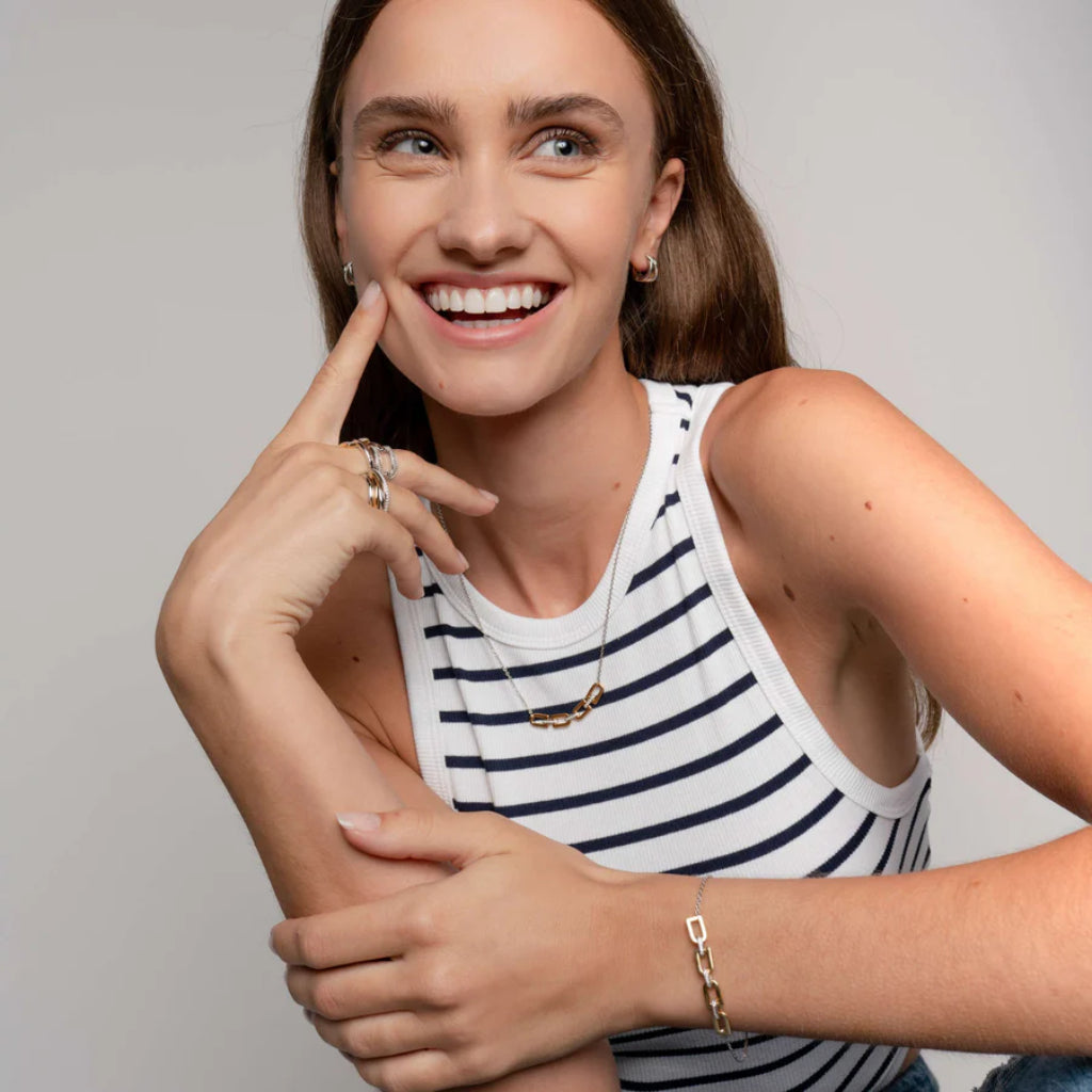  imagem mostra uma mulher sorridente que veste uma blusa de alças listrada com listras brancas e azul marinho. A pessoa também está usando vários anéis nos dedos, uma pulseira no pulso esquerdo e um colar com um pequeno pingente.