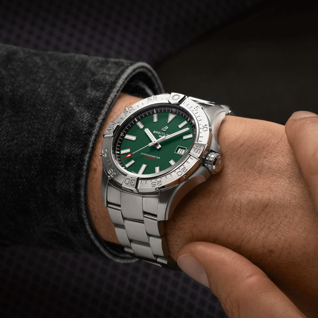 Uma imagem em close-up do pulso de uma pessoa usando um relógio de luxo com uma pulseira de prata e mostrador verde. O relógio tem um indicador de data, marcas de hora brancas, e os ponteiros estão ajustados para 10:10. A moldura tem números gravados e está girada ligeiramente para a esquerda. A pessoa está usando uma manga de cor escura, e o fundo é cinza escuro texturizado.