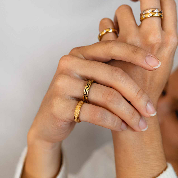 Imagem em close-up das mãos de uma pessoa exibindo vários anéis nos dedos. Os anéis têm designs diferentes, incluindo o que parece ser um padrão de chave grega e outras bandas texturizadas. A pessoa está vestindo uma roupa branca, sugerindo um foco na joalheria em vez do traje.