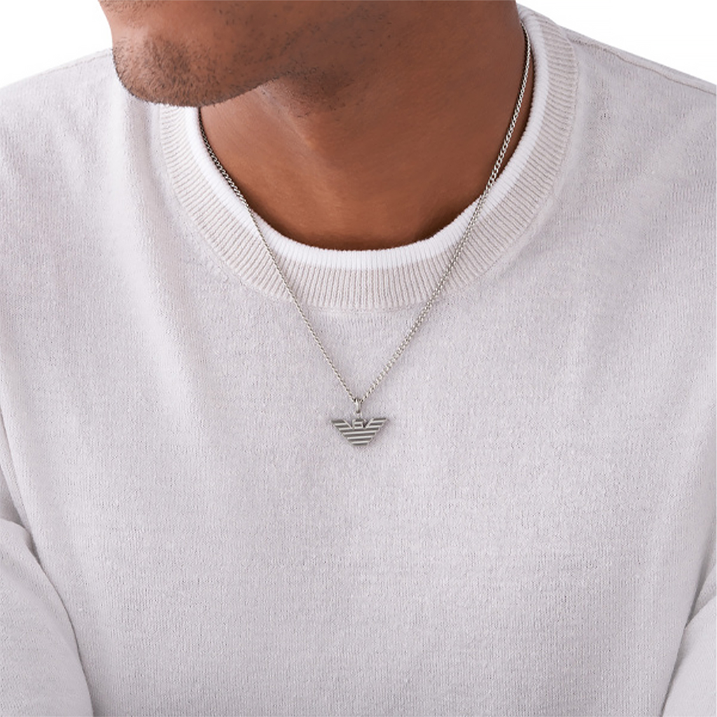 Imagem de um colar de aço inoxidável da Emporio Armani usado por uma pessoa com uma camisola branca de gola em V. O colar apresenta um pingente proeminente com o distintivo logotipo da águia da Emporio Armani.