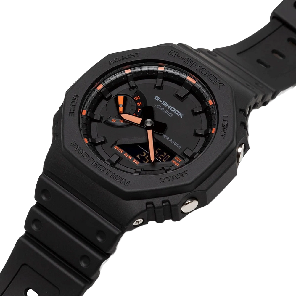Um relógio de pulso G-Shock de resina preta com um mostrador digital-analógico. O mostrador apresenta acentos laranja e brancos nos ponteiros e marcadores de hora. A moldura ao redor do mostrador inclui etiquetas como ‘Adjust’, ‘Mode’, ‘Light’ e ‘Search’. Há também etiquetas para diferentes funções como ‘Shock Resist’ e ‘Water 20BAR Resist’. A pulseira é preta, combinando com o corpo do relógio, e parece ser feita de um material durável, adequado para uso ativo ou em ambientes rigorosos