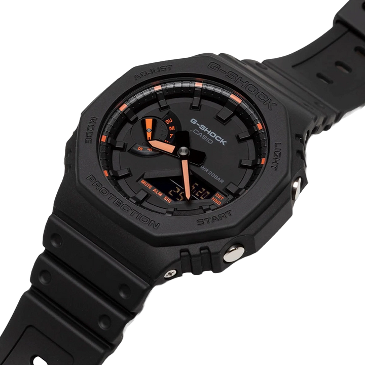 Um relógio de pulso G-Shock de resina preta com um mostrador digital-analógico. O mostrador apresenta acentos laranja e brancos nos ponteiros e marcadores de hora. A moldura ao redor do mostrador inclui etiquetas como ‘Adjust’, ‘Mode’, ‘Light’ e ‘Search’. Há também etiquetas para diferentes funções como ‘Shock Resist’ e ‘Water 20BAR Resist’. A pulseira é preta, combinando com o corpo do relógio, e parece ser feita de um material durável, adequado para uso ativo ou em ambientes rigorosos