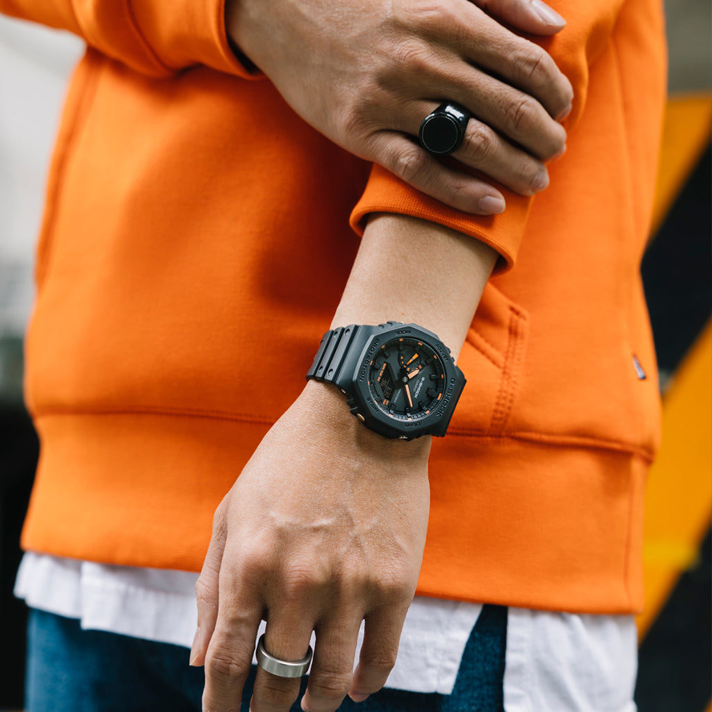 Um relógio de resina de cor escura com mostrador laranja no pulso esquerdo de uma pessoa. A pessoa está vestindo uma blusa de manga comprida laranja e calças brancas, e usa dois anéis nos dedos. O design do relógio é ousado, com marcadores de hora e ponteiros visíveis, destacando-se contra a cor vibrante da blusa.