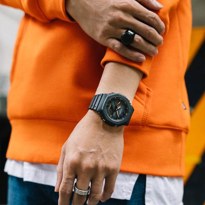 Um relógio de resina de cor escura com mostrador laranja no pulso esquerdo de uma pessoa. A pessoa está vestindo uma blusa de manga comprida laranja e calças brancas, e usa dois anéis nos dedos. O design do relógio é ousado, com marcadores de hora e ponteiros visíveis, destacando-se contra a cor vibrante da blusa.
