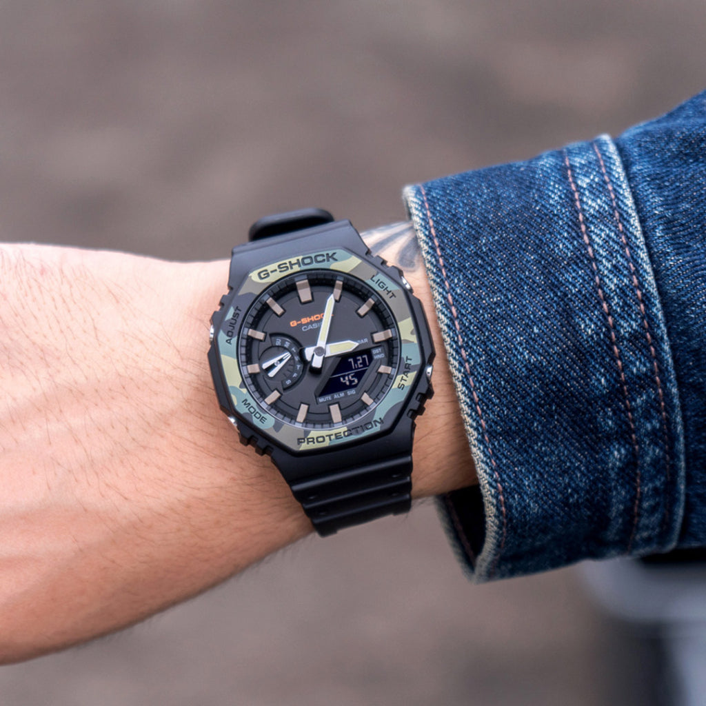 :magem em close-up de um pulso usando um relógio G-SHOCK de resina preta. O relógio possui um mostrador digital-analógico com ponteiros brancos e amarelos, e vários botões na lateral da caixa. A hora no relógio marca 03:05, e há uma leitura digital logo abaixo dos ponteiros mostrando ‘7:27’. O fundo está desfocado, destacando o design e as funcionalidades do relógio.