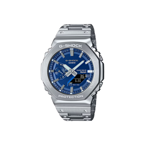 Um relógio Casio G-Shock de aço inoxidável com uma pulseira e caixa prateadas. O mostrador é azul e possui displays digital e analógico mostrando a hora. O relógio tem vários botões nas laterais, um display de dia na parte superior e vários indicadores para alarme, sinal e vida útil da bateria. As palavras “Shock Resist” indicam sua durabilidade, e “Water Resist 20 BAR” sugere uma resistência significativa à água.