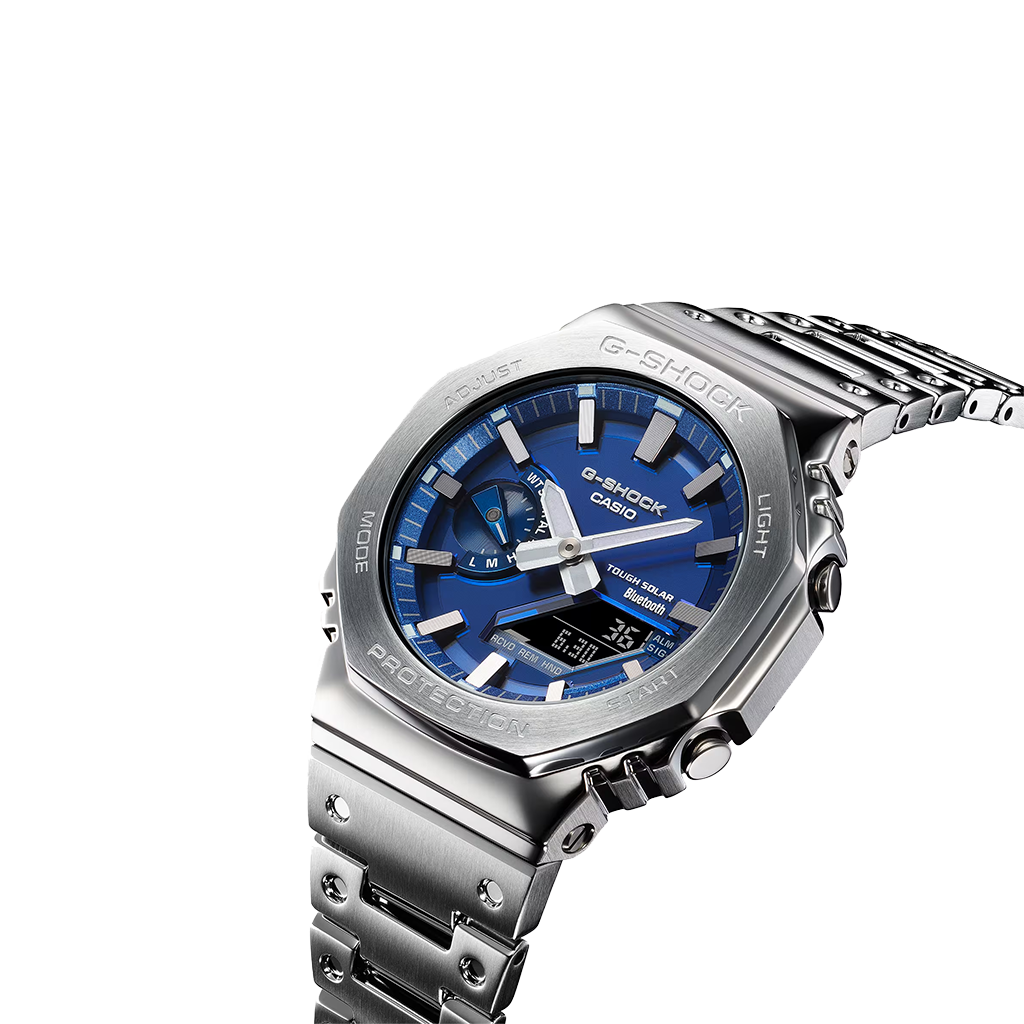 Um relógio Casio G-Shock de aço inoxidável com mostrador analógico azul, marcadores de horas e ponteiros prateados, e um display digital na posição das 6 horas. O relógio tem um design robusto com um aro hexagonal, botões laterais para ajustes e está preso a uma pulseira de aço inoxidável combinando. O fundo é branco, destacando as características do relógio.