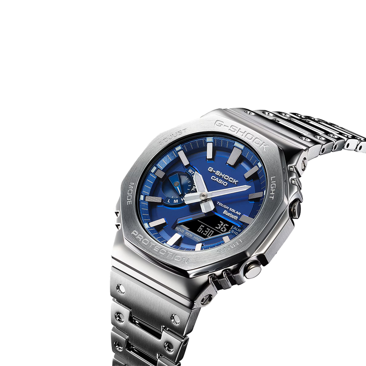 Um relógio Casio G-Shock de aço inoxidável com mostrador analógico azul, marcadores de horas e ponteiros prateados, e um display digital na posição das 6 horas. O relógio tem um design robusto com um aro hexagonal, botões laterais para ajustes e está preso a uma pulseira de aço inoxidável combinando. O fundo é branco, destacando as características do relógio.