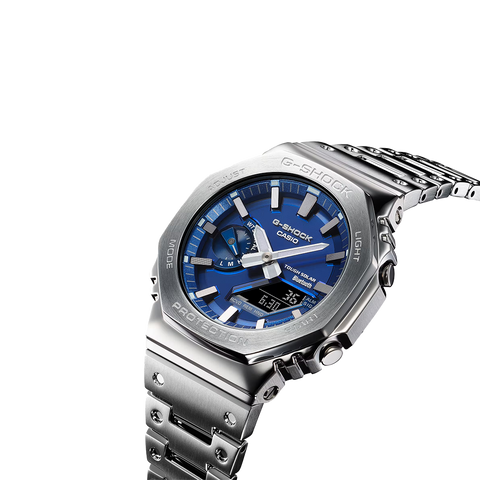 Um relógio Casio G-Shock de aço inoxidável com mostrador analógico azul, marcadores de horas e ponteiros prateados, e um display digital na posição das 6 horas. O relógio tem um design robusto com um aro hexagonal, botões laterais para ajustes e está preso a uma pulseira de aço inoxidável combinando. O fundo é branco, destacando as características do relógio.