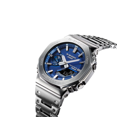 Um relógio Casio G-Shock de aço inoxidável com mostrador analógico azul, marcadores de horas e ponteiros prateados, e um display digital na posição das 6 horas. O relógio tem um design robusto com um aro hexagonal, botões laterais para ajustes e está preso a uma pulseira de aço inoxidável combinando. O fundo é branco, destacando as características do relógio.