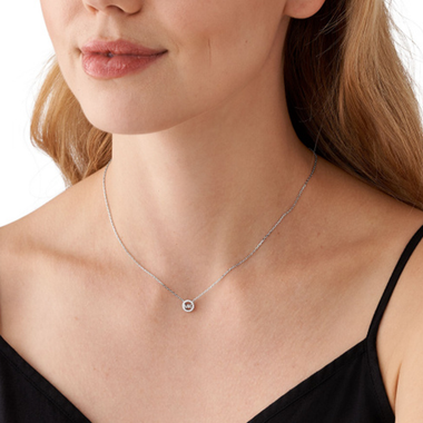 Colar de prata banhada a ródio com um pingente de zircónia, exibido em uma pessoa vestindo uma blusa preta. A corrente delicada sustenta uma pequena pedra de zircónia redonda e brilhante no centro, oferecendo uma peça de joalheria simples, mas elegante, que se destaca contra o fundo escuro da roupa.