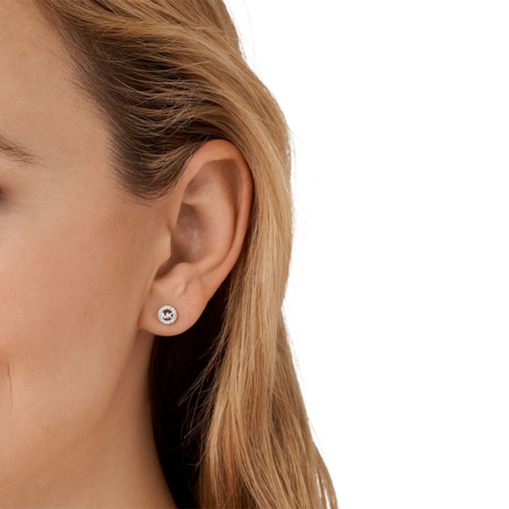 Uma imagem em close-up da orelha de uma pessoa usando um brinco da Michael Kors. O brinco é feito de prata banhada a ródio e apresenta uma pedra de zircónia redonda no centro, dando-lhe um brilho sutil. O design é simples, mas elegante, adequado tanto para uso casual quanto formal.