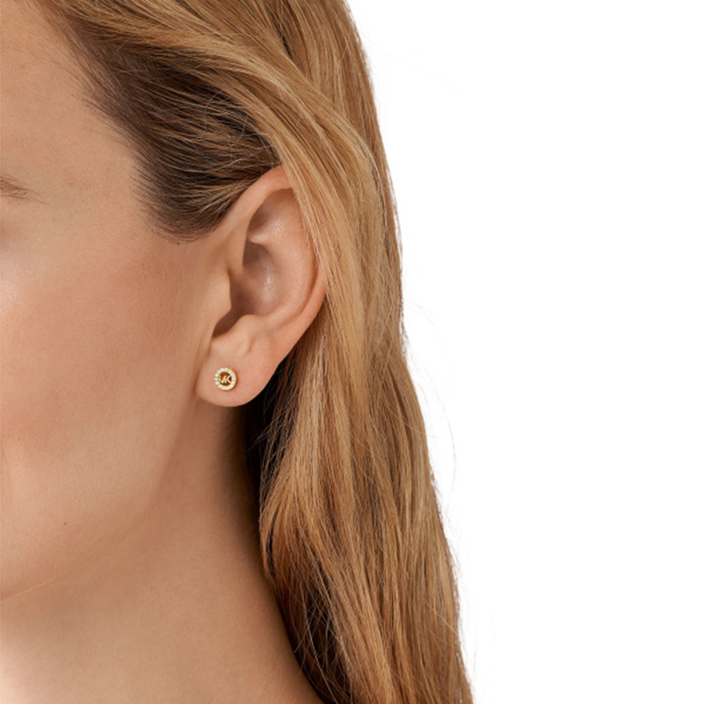 Uma imagem em close-up da orelha de uma pessoa usando um brinco da Michael Kors. O brinco é feito de prata banhada a ouro e apresenta uma pedra de zircônia redonda no centro, dando-lhe um brilho sutil. O design é simples, mas elegante, adequado tanto para uso casual quanto formal.