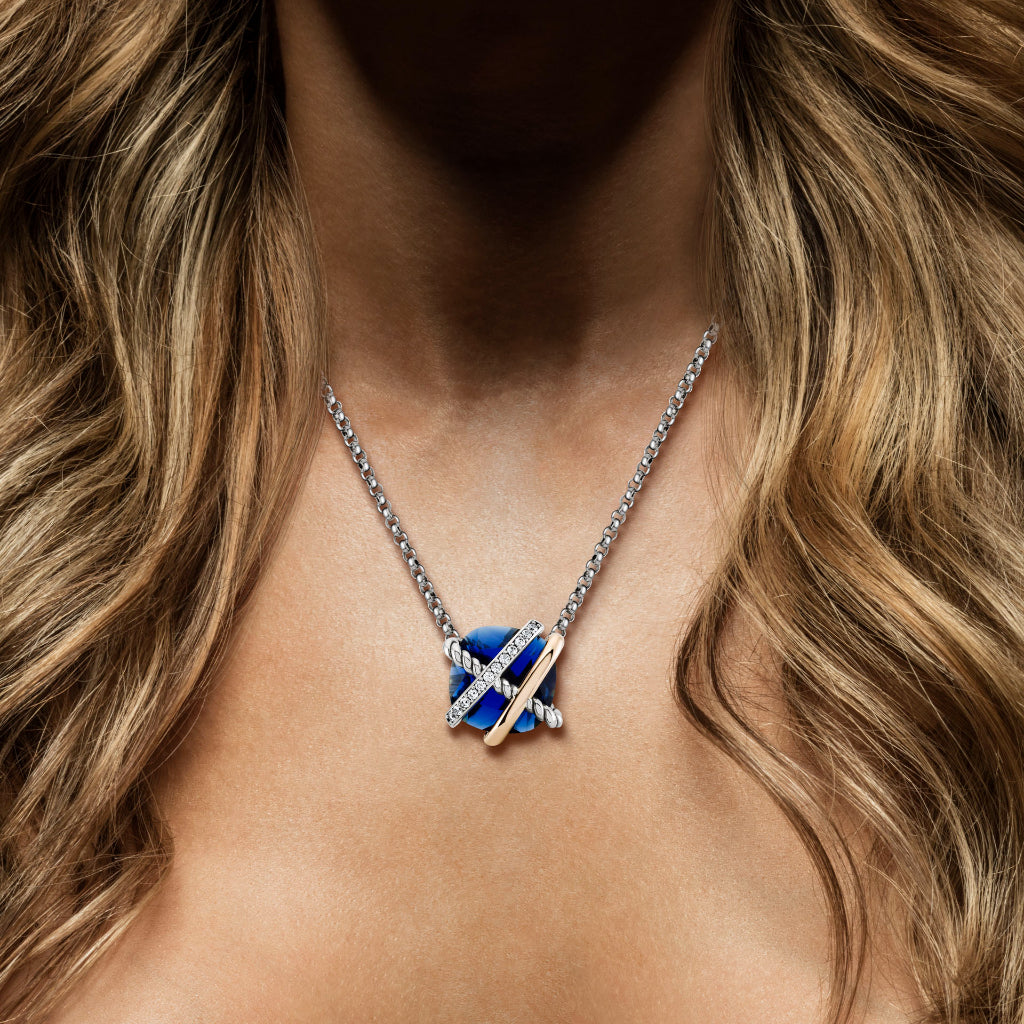 Um colar de prata e ouro com um pingente de zircónia azul. O pingente apresenta um design intrincado com bandas de prata cruzando uma estrutura de ouro, embalando a zircónia azul em forma oval no centro. O colar é exibido contra um fundo de cabelo castanho claro ondulado que desce, o que acentua a elegância da joia e o contraste de cores vibrantes.