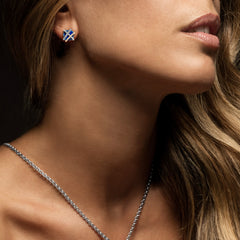 A imagem mostra um close da área do pescoço e ombro de uma mulher. A pessoa está usando um brinco de diamante com um design que se assemelha a uma cruz diagonal branca em um campo azul. Além disso, há um vislumbre do que parece ser um colar de diamantes combinando com o estilo do brinco. O fundo desfocado não fornece contexto adicional. Esta imagem pode ser relevante para tópicos relacionados ao design de joias ou itens de moda inspirados em formas geométricas.