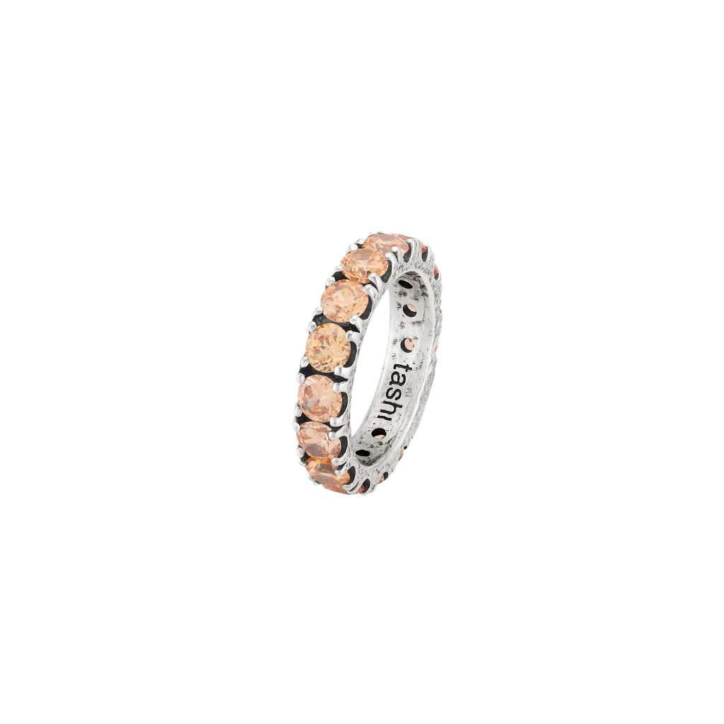 Uma imagem de um anel com uma banda prateada e várias zircónias em tons de pêssego inseridas nela. As zircónias são cortadas em formato redondo e parecem estar espaçadas uniformemente ao redor da banda. O interior da banda tem a marca Tashi gravada