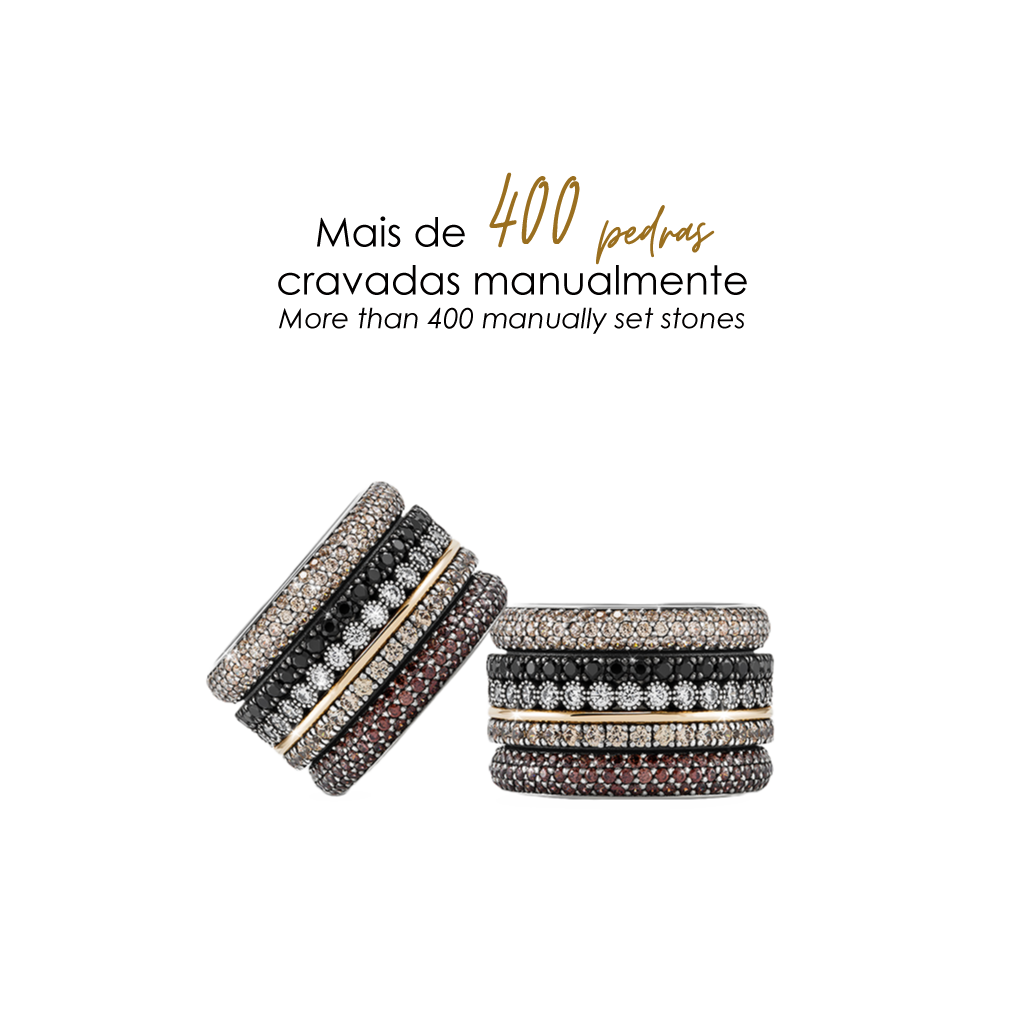 Imagem de dois anéis empilhados com uma variedade de pedras. O anel superior tem três bandas; a banda central é adornada com pedras pretas, ladeada por bandas com pedras brancas e marrons. O anel inferior também tem três bandas, com a central apresentando pedras brancas e as bandas externas exibindo pedras pretas e marrons. Acima dos anéis, o texto em português ‘Mais de 400 pedras cravadas manualmente’ indica o artesanato envolvido na fixação das pedras nestes anéis.