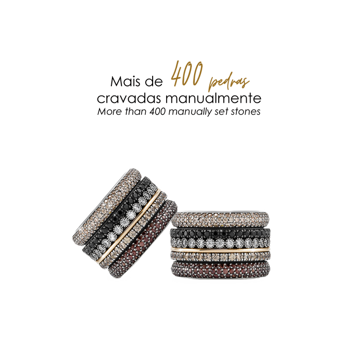 Imagem de dois anéis empilhados com uma variedade de pedras. O anel superior tem três bandas; a banda central é adornada com pedras pretas, ladeada por bandas com pedras brancas e marrons. O anel inferior também tem três bandas, com a central apresentando pedras brancas e as bandas externas exibindo pedras pretas e marrons. Acima dos anéis, o texto em português ‘Mais de 400 pedras cravadas manualmente’ indica o artesanato envolvido na fixação das pedras nestes anéis.