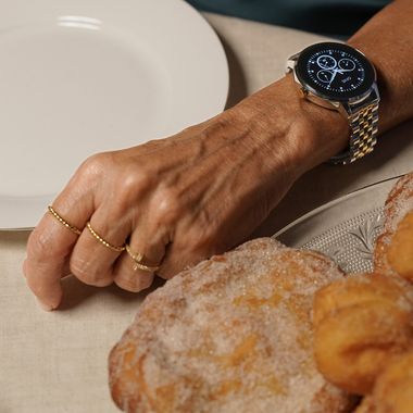Imagem em close-up do pulso de uma pessoa usando um relógio de ouro com mostrador preto e display analógico. O pulso está apoiado em uma mesa perto de um prato branco e alguns doces com açúcar de confeiteiro por cima, sugerindo que o cenário pode ser durante uma refeição ou lanche. A pessoa também usa dois anéis finos de ouro, um no dedo anelar e um no dedo médio.