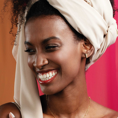 Imagem de mulher a usar um turbante branco e um brinco é visível na orelha. O fundo consiste em duas cores distintas, rosa de um lado e um tom mais escuro, possivelmente castanho ou bordô, do outro lado.