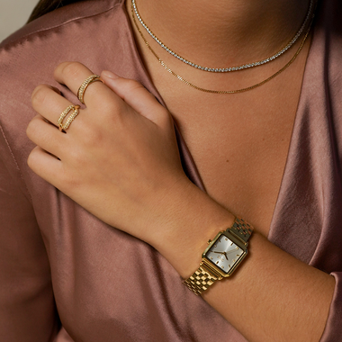 Close-up do pulso esquerdo de uma pessoa usando um relógio dourado com uma face branca quadrada e numerais romanos. A pessoa também está usando duas finas correntes de ouro e um anel de ouro correspondente no dedo indicador, tudo contra uma blusa rosa cetim.