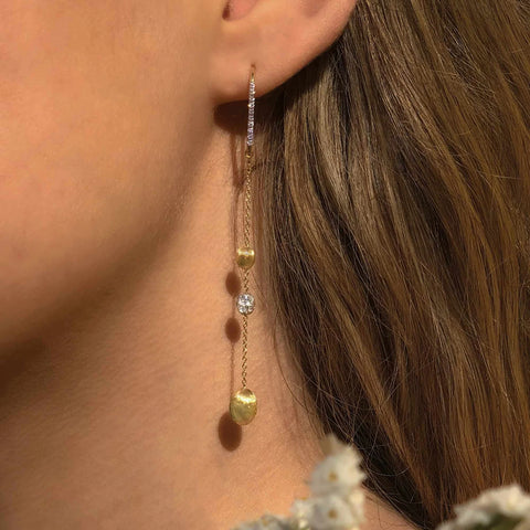 Senhora com os Brincos NANIS Soffio Long dourados com fileira em zircónias no topo, seguido de fio, intercalado com pendentes ovais lisos de tamanhos diferentes e no meio outros pendentes ovais revestidos com zircónias