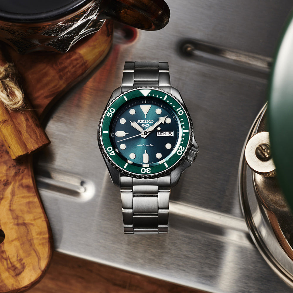 Relógio Seiko 5 Sports SKX Sports Style 42.5mm prateado com mostrador verde e indicação de semana e dia do mês pousado em superficie de metal