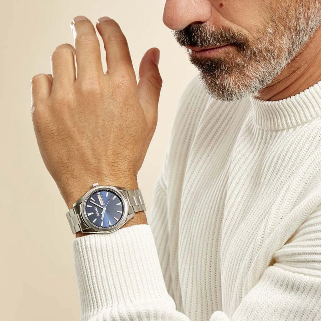 Pulso de uma pessoa usando um relógio de aço inoxidável com mostrador azul e ponteiros prateados. O relógio tem uma pulseira metálica elegante e está posicionado sobre o punho de um suéter branco. A mão da pessoa está ligeiramente levantada contra um fundo neutro, destacando a elegância do relógio.