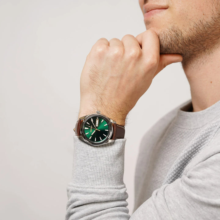 Relógio de pulso com pulseira de couro bovino marrom, exibido no pulso de uma pessoa. O mostrador é verde com múltiplos marcadores e ponteiros, envolto em uma moldura metálica marrom. A pessoa está vestindo uma camisa de manga comprida cinza claro e posando com a mão perto do queixo, destacando o relógio contra um fundo neutro.