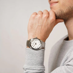 Um relógio de pulso com caixa e pulseira de aço inoxidável. O mostrador é branco com marcadores de hora prateados e ponteiros finos. O design é simples e elegante, adequado para uso diário e ocasiões formais.