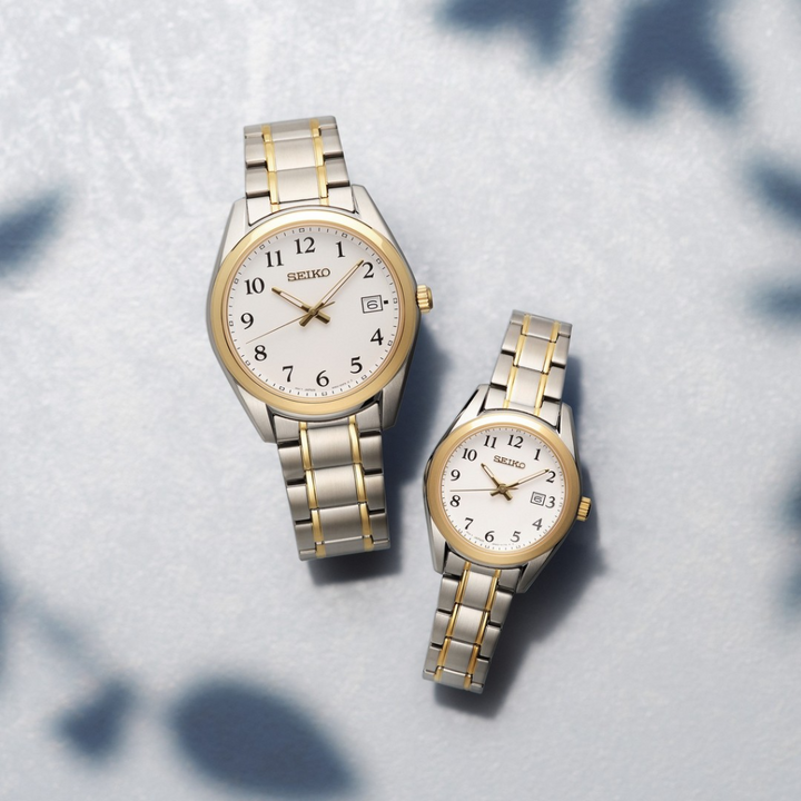 Dois relógios de aço inoxidável Seiko com detalhes dourados, dispostos sobre uma superfície com uma leve camada de neve. O relógio à esquerda é maior e possui um indicador de data na posição das 3 horas, enquanto o da direita é menor e sem indicador de data. Ambos têm pulseiras metálicas, mostradores brancos com algarismos arábicos pretos para cada hora e ponteiros dourados. O logotipo da Seiko está visível abaixo da posição das 12 horas em ambos os mostradores.