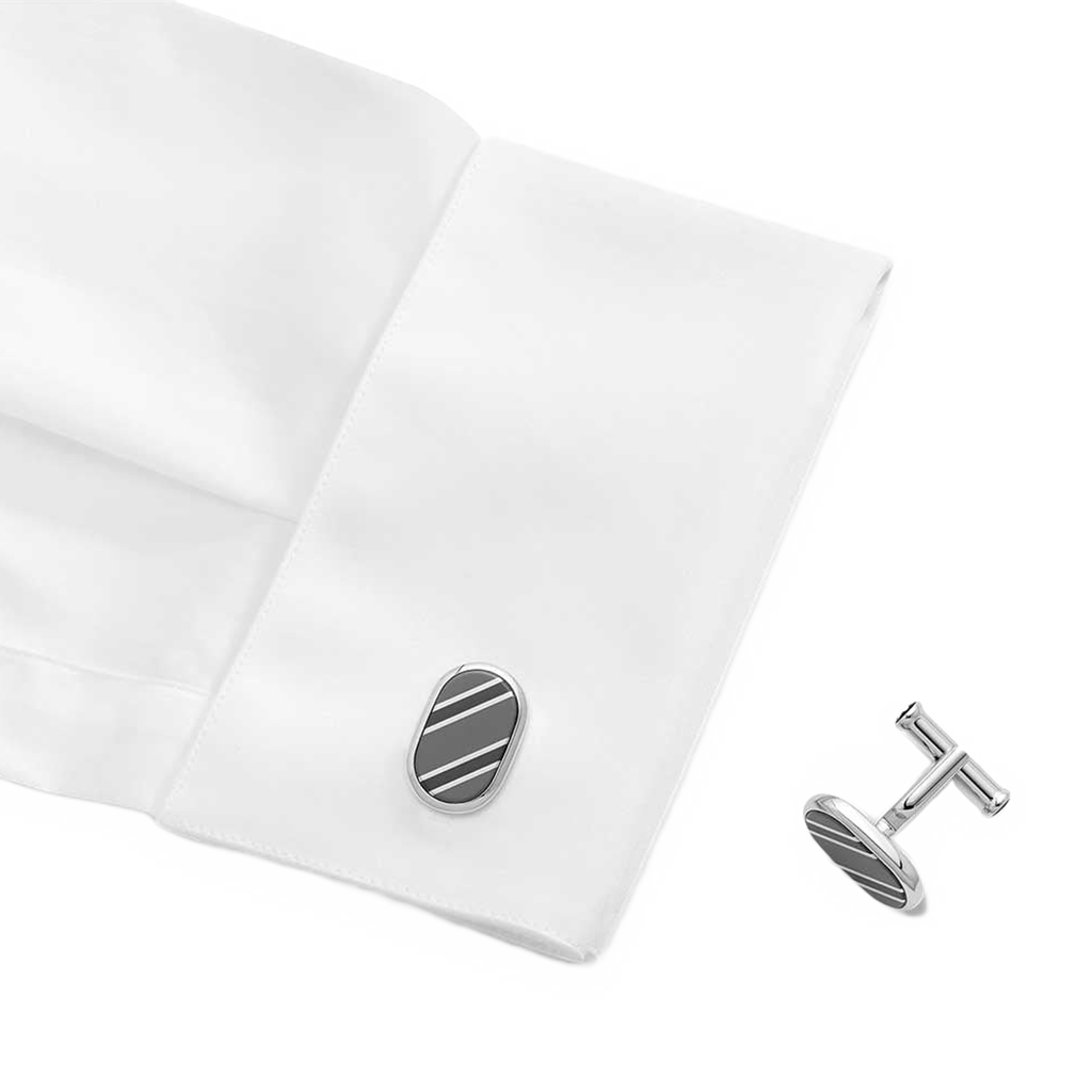 Um par de botões de punho em formato oval com acabamento em prata e um design de uma única listra preta, exibidos no punho de uma camisa social branca. Os botões de punho são elegantes e proporcionam um acessório sutil, mas sofisticado, para o traje formal.