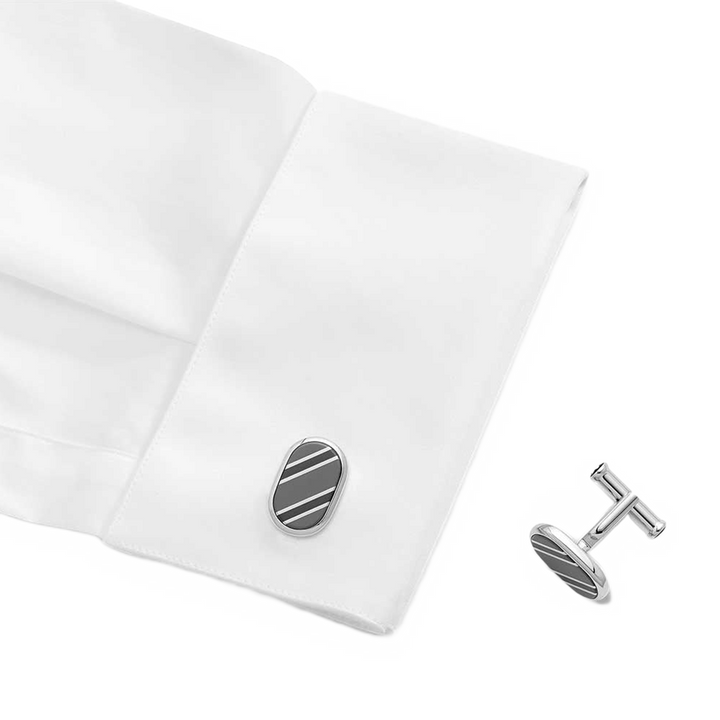 Um par de botões de punho em formato oval com acabamento em prata e um design de uma única listra preta, exibidos no punho de uma camisa social branca. Os botões de punho são elegantes e proporcionam um acessório sutil, mas sofisticado, para o traje formal.