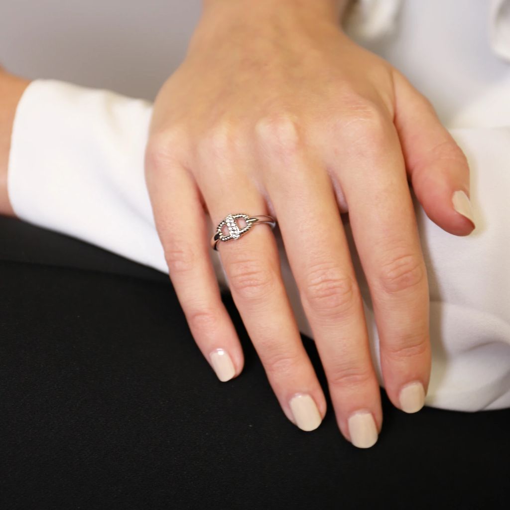 A imagem mostra um close-up de uma mão repousando sobre uma superfície plana, provavelmente pertencente a um adulto. A mão está adornada com um anel que apresenta uma grande pedra central ladeada por pedras menores em uma banda de prata ou ouro branco. As unhas estão manicuradas com esmalte pálido, e a pessoa está vestindo uma manga branca, sugerindo um elemento de formalidade ou elegância. Não há problemas de matemática ou dever de casa presentes na imagem para transcrever.