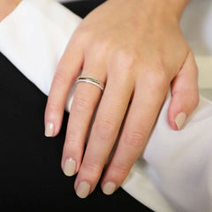 Uma imagem em close-up de uma mão repousando sobre uma superfície branca, exibindo um anel com múltiplos pequenos diamantes incrustados em uma banda no dedo anelar. A mão tem unhas bem cuidadas com esmalte claro.