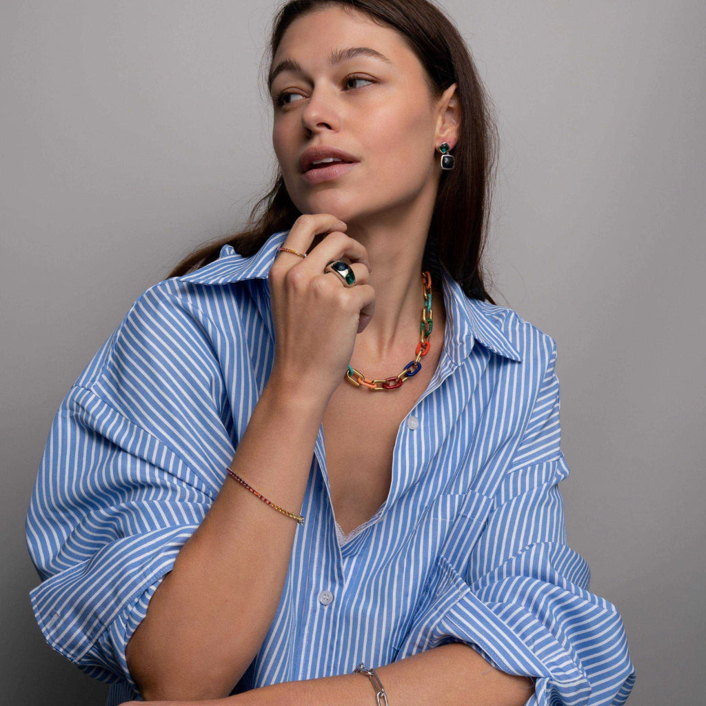 Imagem de um indivíduo do peito para cima, vestindo uma camisa listrada de azul e branco com os dois primeiros botões desabotoados. O rosto do indivíduo está obscurecido por uma área pixelizada marrom, mantendo a privacidade. A pessoa está segurando uma caneta ou lápis verde perto do queixo com a mão esquerda, que tem as unhas pintadas. Os acessórios visíveis incluem um par de brincos pendentes e um colar de contas coloridas.