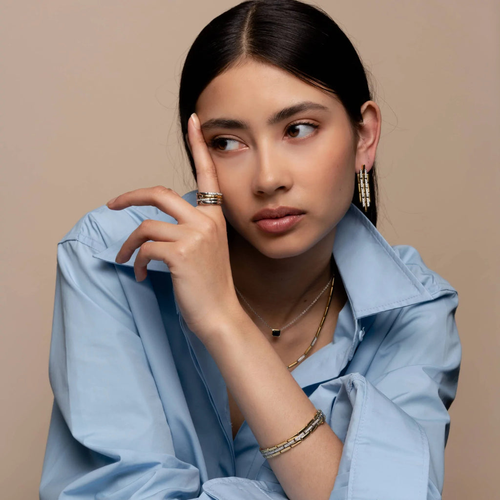 imagem de mulher de cabelo escuro, com argolas largas nas orelhas, o dedo indicador levantado e com um anel , vários colares ao pescoço e uma pulseira no braço.