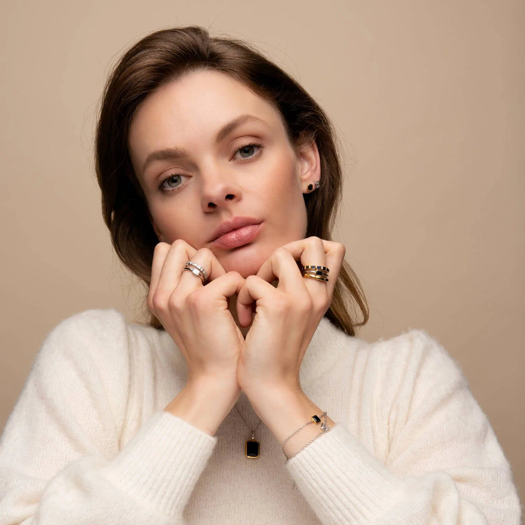 imagem de mulher de cabelo castanho escuro a encarar a câmara. A mulher tem o queixo pousado nas mãos e ambos os dedos do meio adornados com aneis. No pulso esquerdo tem uma fina pulseira. Veste uma camisola de lã branca e é possível ver um colar com pingente ao peito