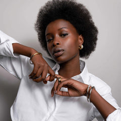 Imagem de uma pessoa vestindo uma camisa branca e joias douradas, incluindo brincos e anéis. O indivíduo está formando um coração com as mãos. O rosto está obscurecido por pixelização para privacidade.