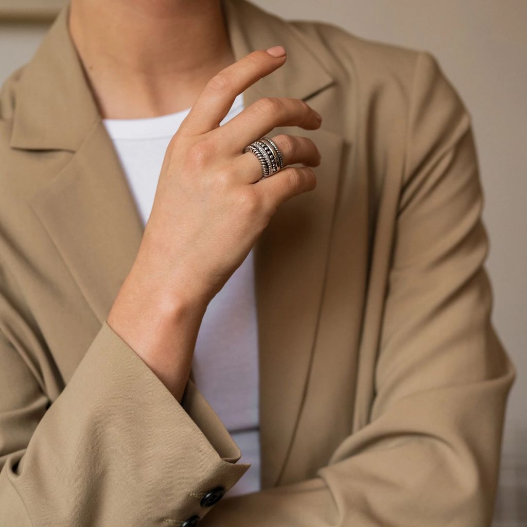 A imagem mostra um close-up do tronco e da mão de uma pessoa. A pessoa está vestindo um blazer bege sobre uma blusa branca. A mão está fazendo um gesto de ‘OK’ com o polegar e o indicador tocando para formar um círculo, enquanto os dedos restantes estão estendidos para cima. No dedo anelar, há um anel de prata com gravações detalhadas. O fundo é neutro e indescritível, concentrando a atenção no gesto e no anel.