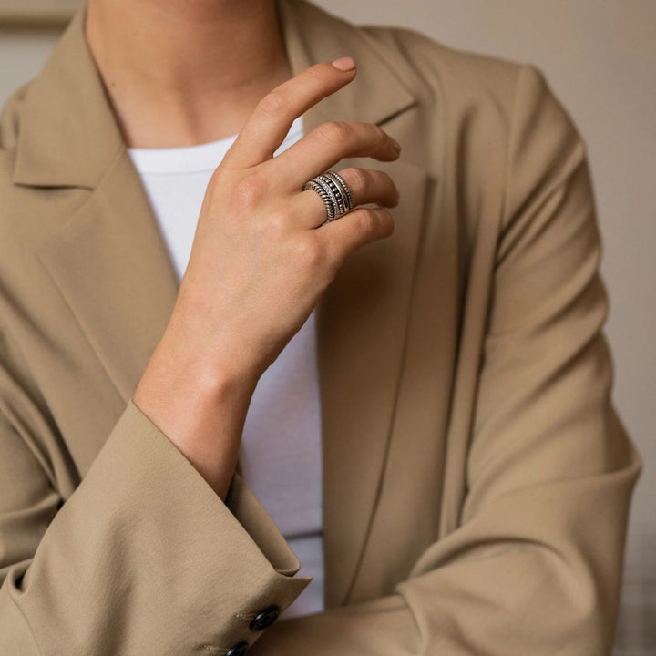 A imagem mostra um close-up do tronco e da mão de uma pessoa. A pessoa está vestindo um blazer bege sobre uma blusa branca. A mão está fazendo um gesto de ‘OK’ com o polegar e o indicador tocando para formar um círculo, enquanto os dedos restantes estão estendidos para cima. No dedo anelar, há um anel de prata com gravações detalhadas. O fundo é neutro e indescritível, concentrando a atenção no gesto e no anel.