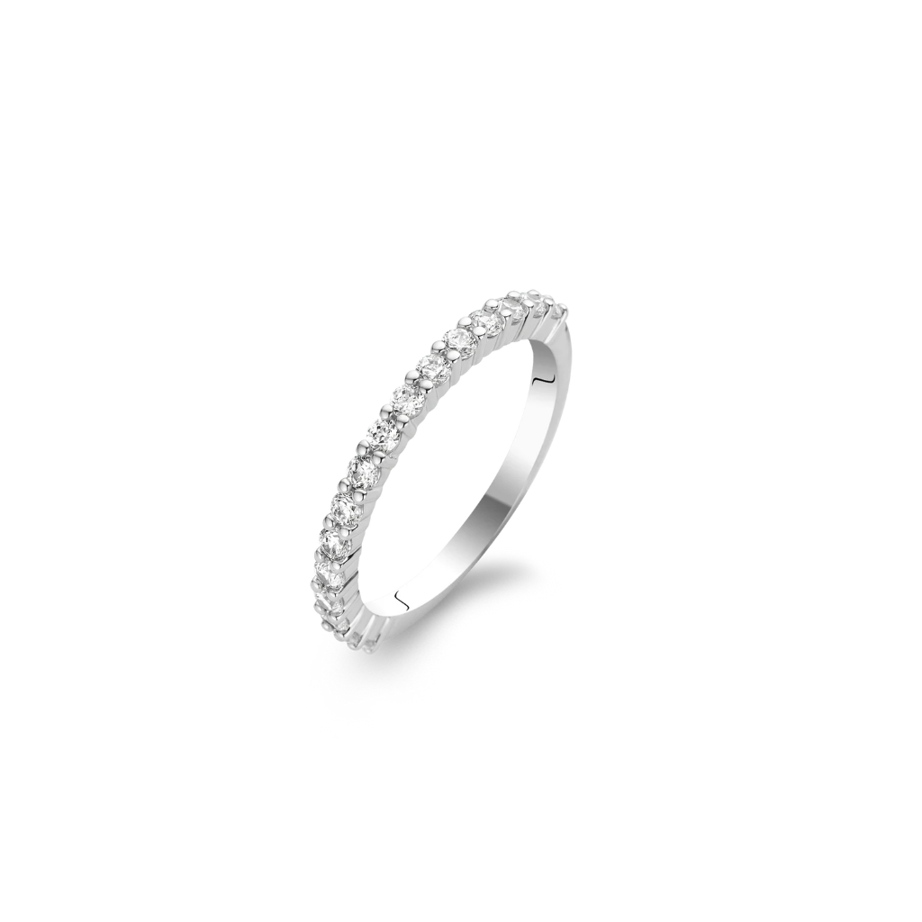 Imagem de um anel em aço banhado a platina, adornado com várias pedras de zircónia de corte redondo, dispostas em uma única fileira ao longo da metade superior da banda. O anel é exibido contra um fundo branco simples, destacando seu acabamento lustroso e a clareza cintilante das gemas.