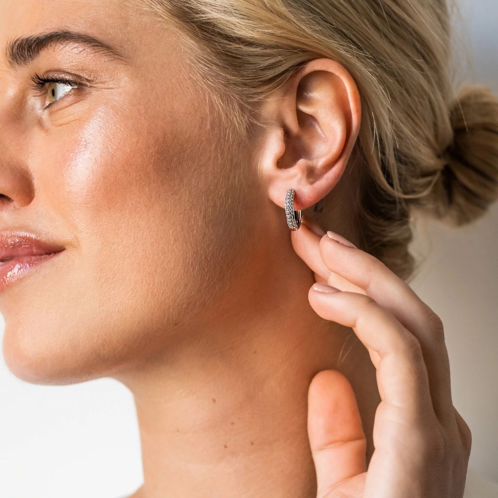 Close-up do perfil lateral de uma mulher sorridente,  mostrando uma orelha adornada com uma argola prateada com zircónias . A pessoa tem o cabelo preso em um coque, e os dedos estão tocando suavemente o brinco. O fundo está desfocado para focar no brinco e na orelha.
